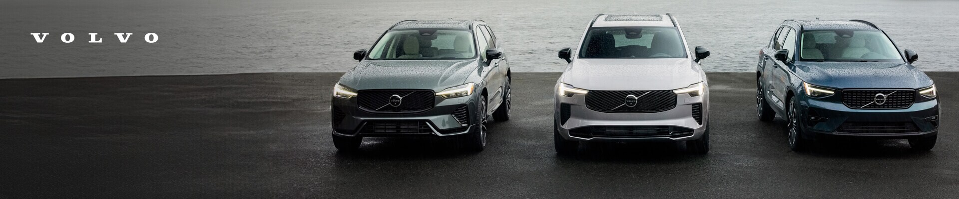 Front view of grey, silver, and blue Volvo SUVs lined up on wet asphalt in front of a river.