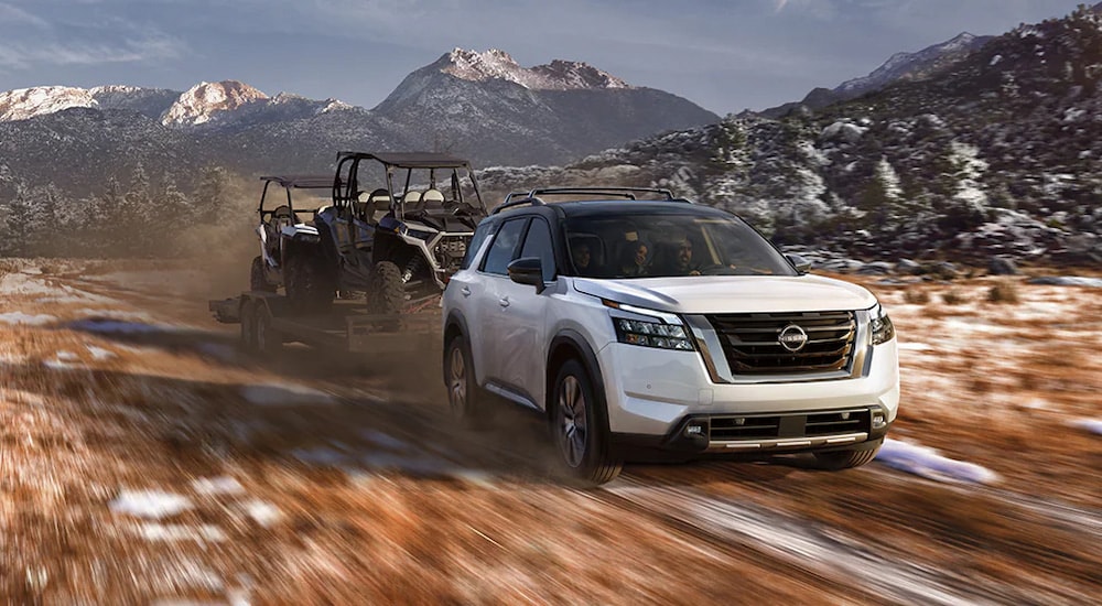 A white 2025 Nissan Pathfinder Platinum towing ATVs on a trailer through the mountains.
