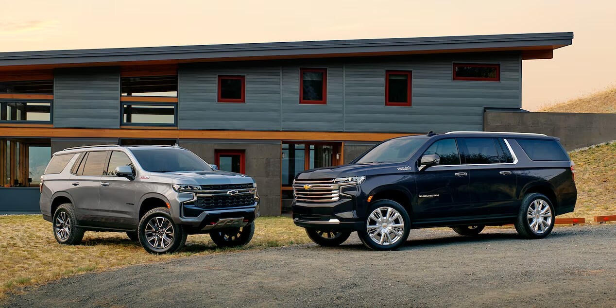 Two Chevrolet Suburban SUVs parked in front of a modern house. Sterling Gray Metallic on the left, Midnight Blue Metallic on the right.