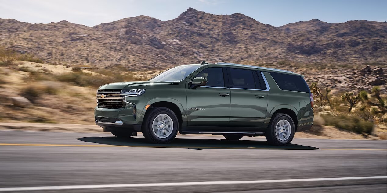 A Silver Sage Metallic Chevrolet Suburban drives on a road through a desert landscape with mountains in the background under a clear blue sky.