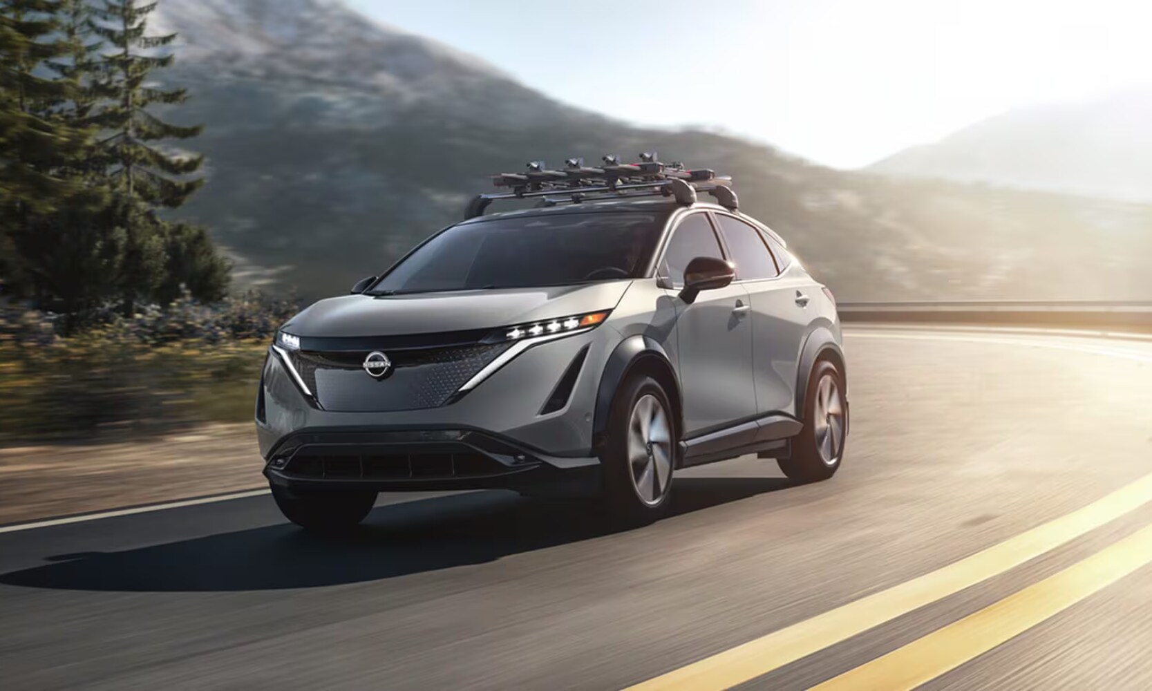 2026%20Nissan%20ARIYA%20Towing%20Capcity%20%20Nissan%20of%20Silsbee%20TX.png Exterior front-view image of a silver 2026 Nissan ARIYA driving on a freeway with a roof-mounted bike rack