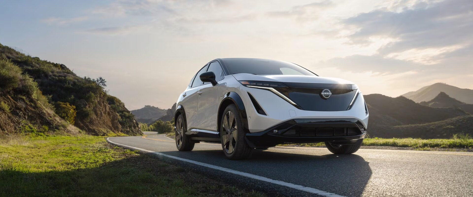 Exterior front-view image of a white 2026 Nissan ARIYA driving on an open mountainous freeway