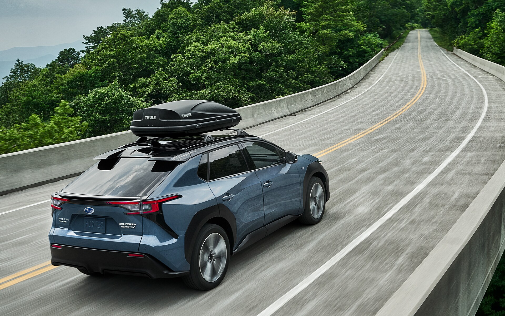 A Subaru Solterra EV with a Thule cargo carrier drives over a high bridge over a wooded gorge.