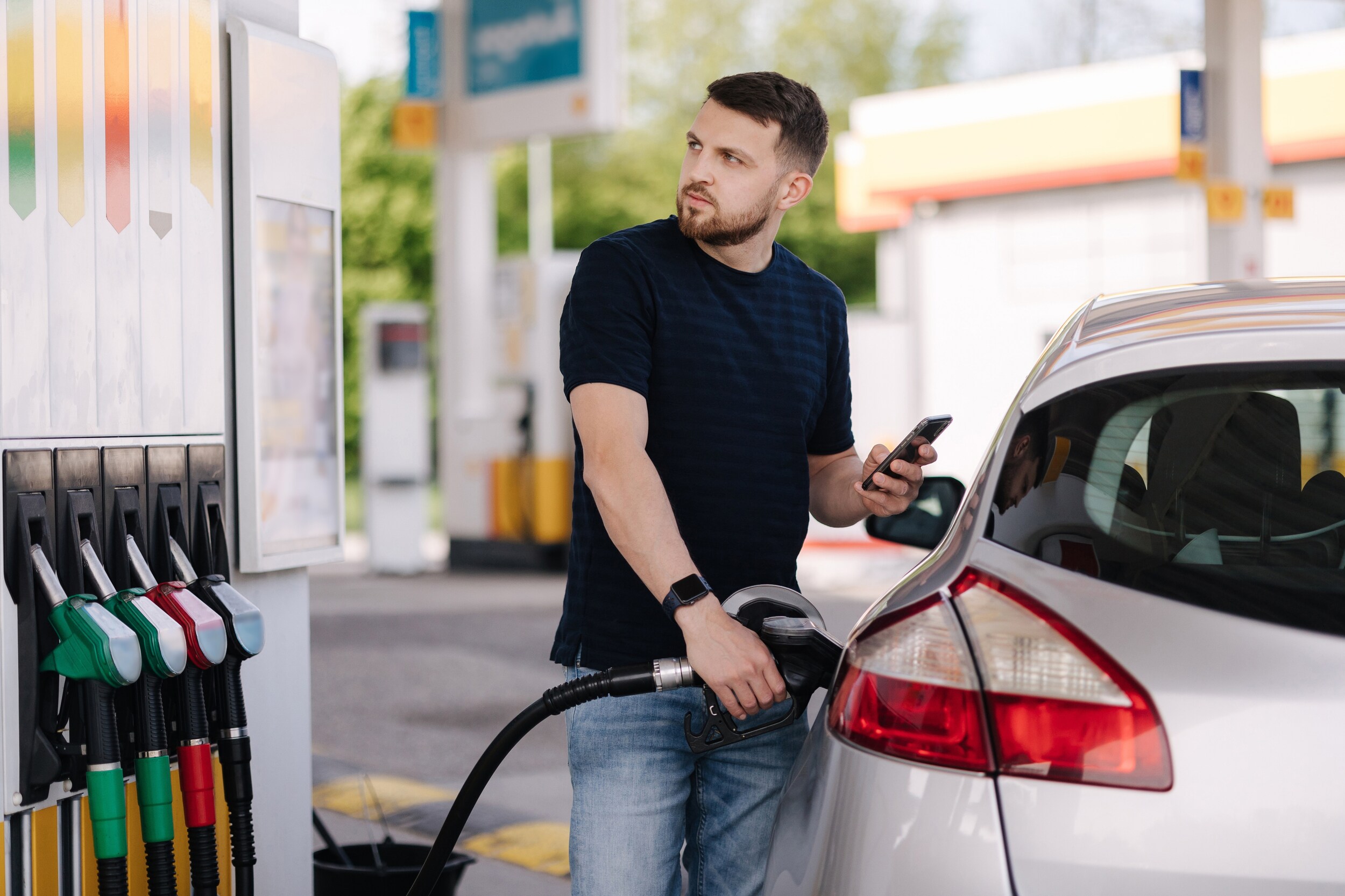 A person pumping gas into a car while holding a phone and looking at the fuel pump station A person pumping gas into a car while holding a phone and looking at the fuel pump station