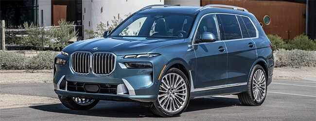 Front three-quarter view of a blue BMW X7 parked in a stylish desert-like environment with modern architecture