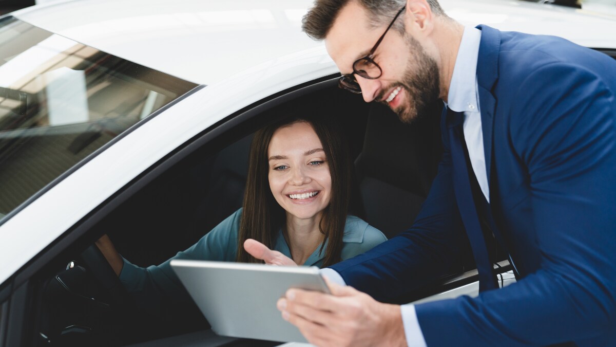 A car salesperson showing a tablet screen to a car buyer, who's behind the wheel of a car A car salesperson showing a tablet screen to a car buyer, who's behind the wheel of a car