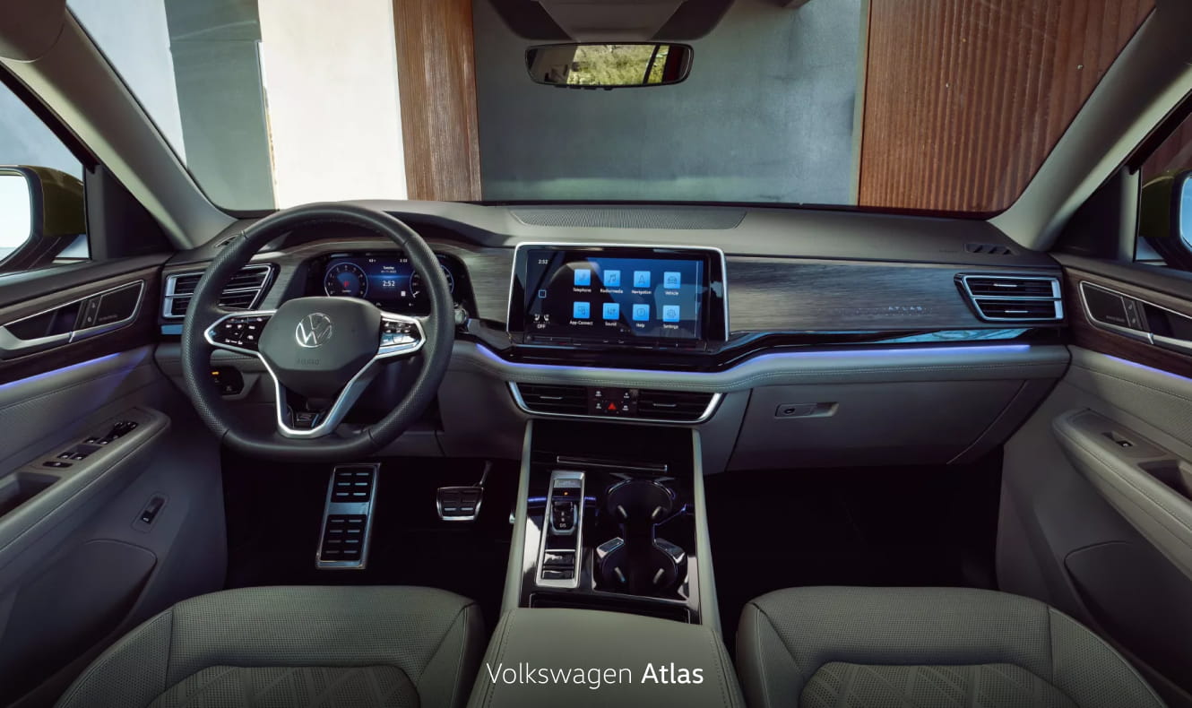 Volkswagen Atlas interior featuring a digital driver display, touchscreen infotainment system, and ambient lighting with a modern architectural setting visible through the windshield.