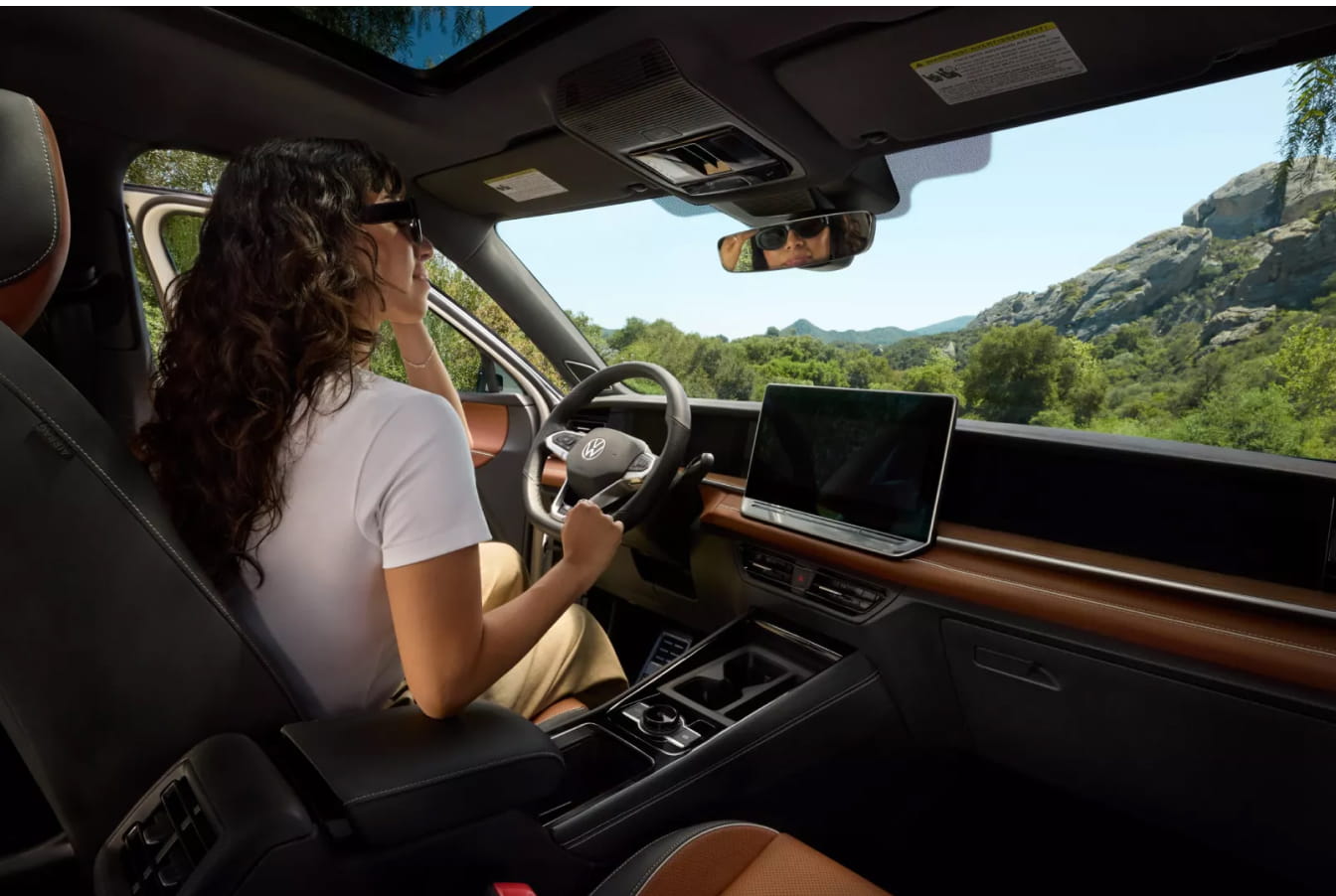 A woman drives a Volkswagen Tiguan SUV through a scenic area with green hills and rocks. She wears sunglasses and looks content, highlighting road trip leisure.
