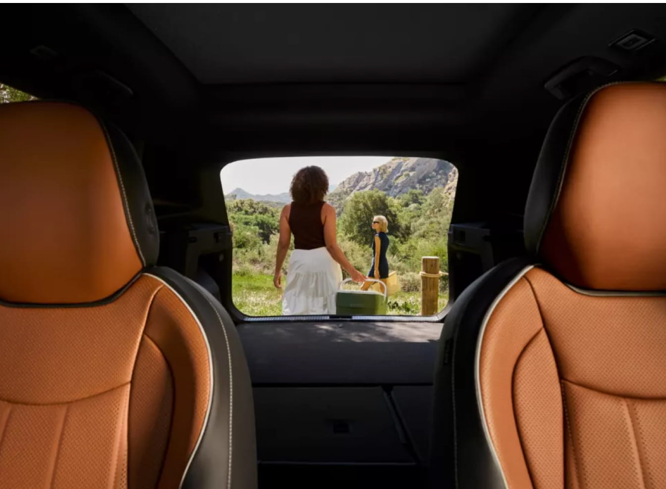 View from inside a Volkswagen Tiguan shows two people walking in a lush landscape, carrying a picnic basket. The car has tan leather seats, conveying a relaxed, adventurous feel.