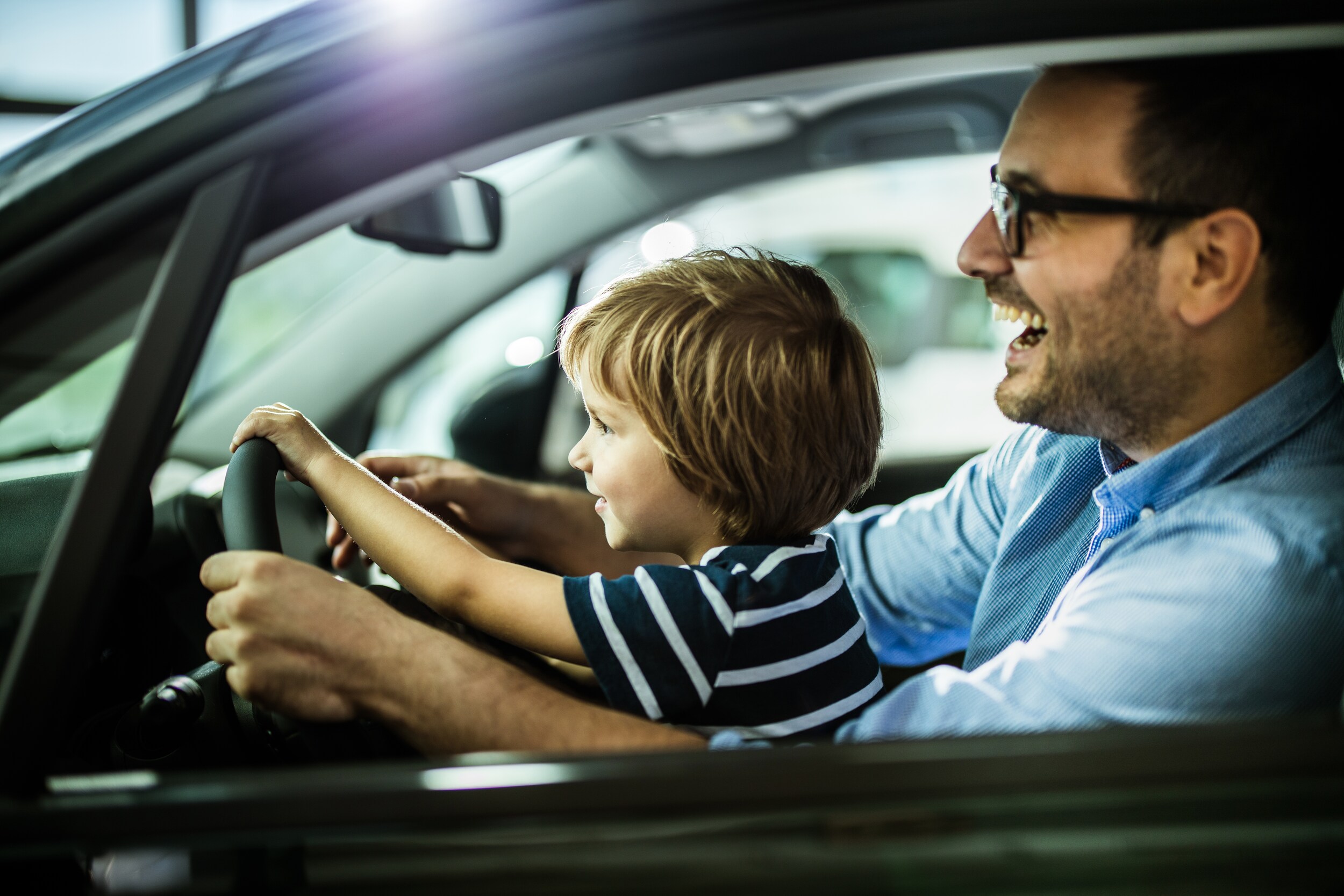 used-vs-new-car.jpg a young boy sitting on his fathers lap in the drivers seat pretending to drive