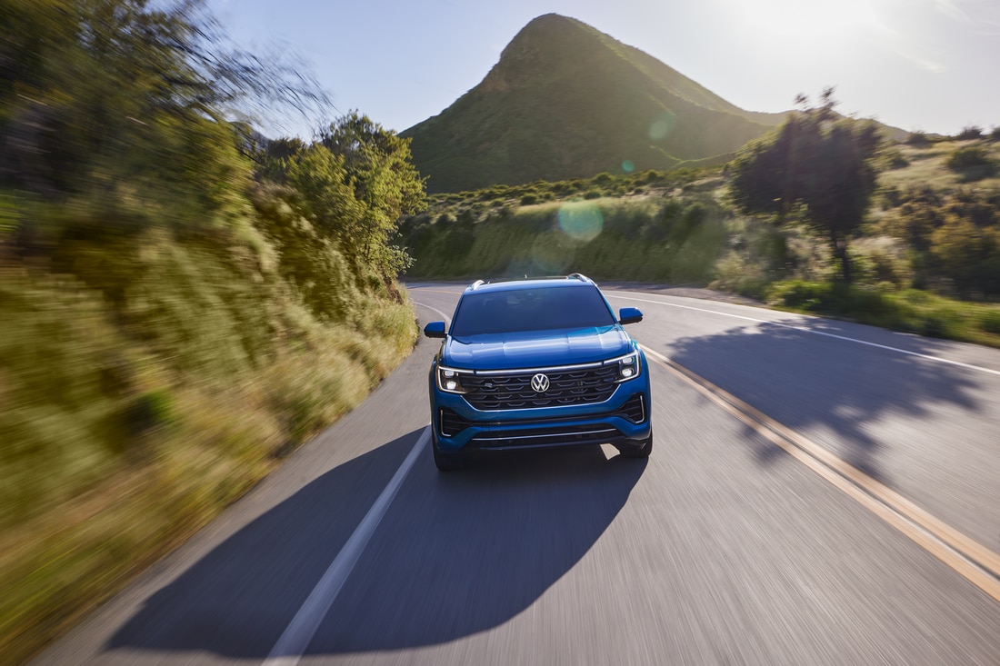 2024-Volkswagen-AtlasCrossSport-SEL-SUV-L02.png blue Volkswagen Atlas Cross Sport SUV driving down the road with a large hill in the background