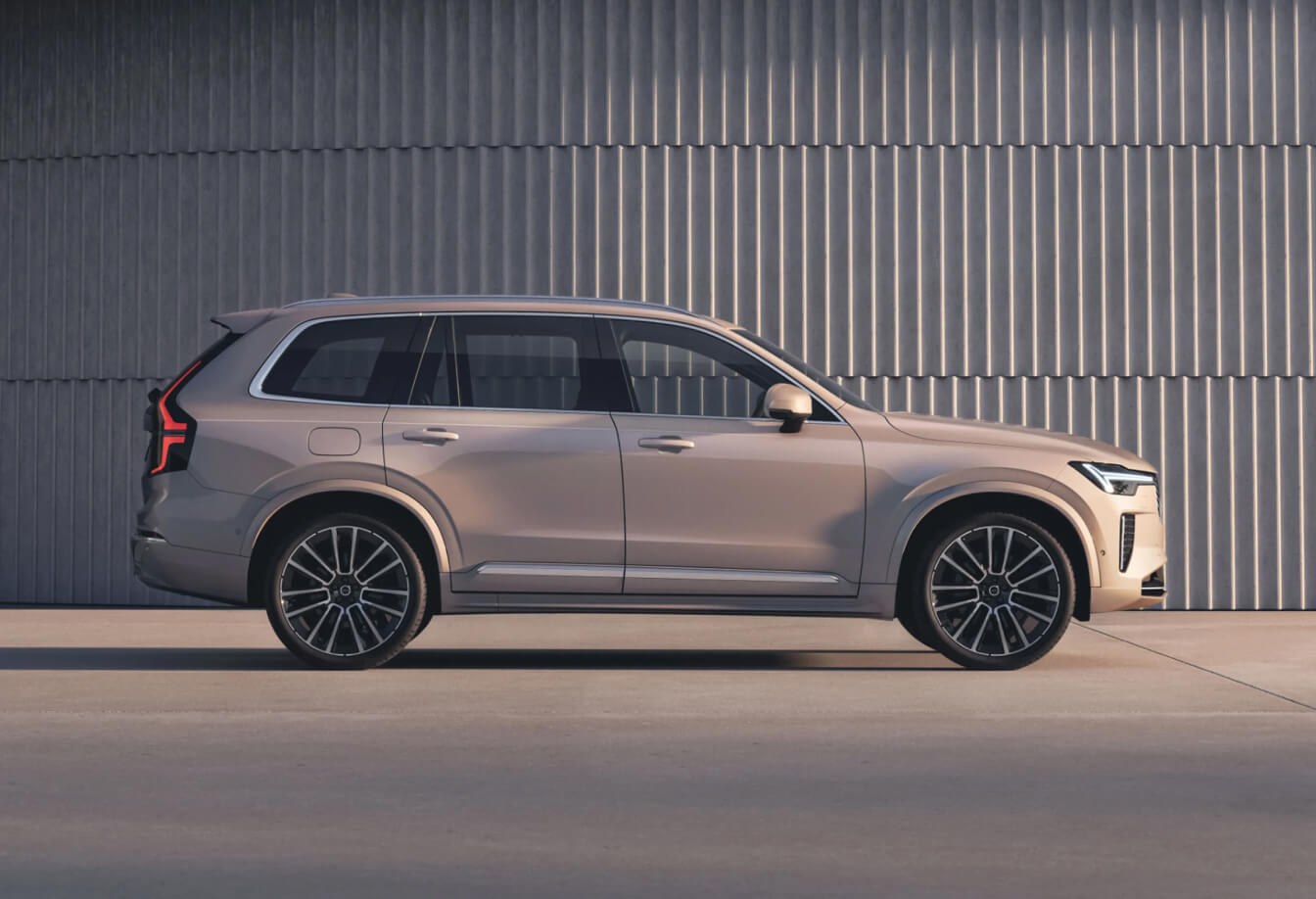Side view of a tan 2026 Volvo XC90 parked in an open lot near a concrete wall.