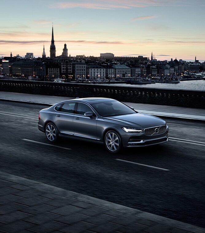 Volvo Cars New Bern is a Car Dealership near River Bend NC | Silver S90 Driving on Road Overlooking City at Sunset Volvo Cars New Bern is a Car Dealership near River Bend NC | Silver S90 Driving on Road Overlooking City at Sunset