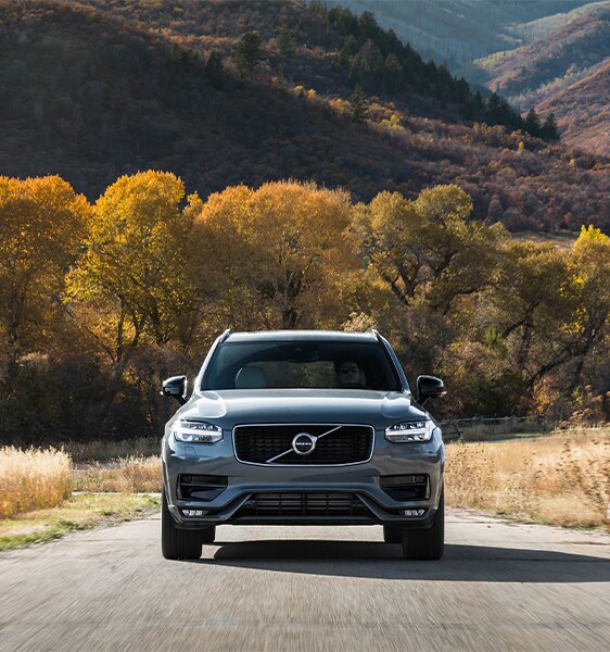 Volvo Cars New Bern is a Car Dealership near Pollocksville, NC | Silver 2020 Volvo XC90 Driving Down Country Road with Yellow Trees in Background