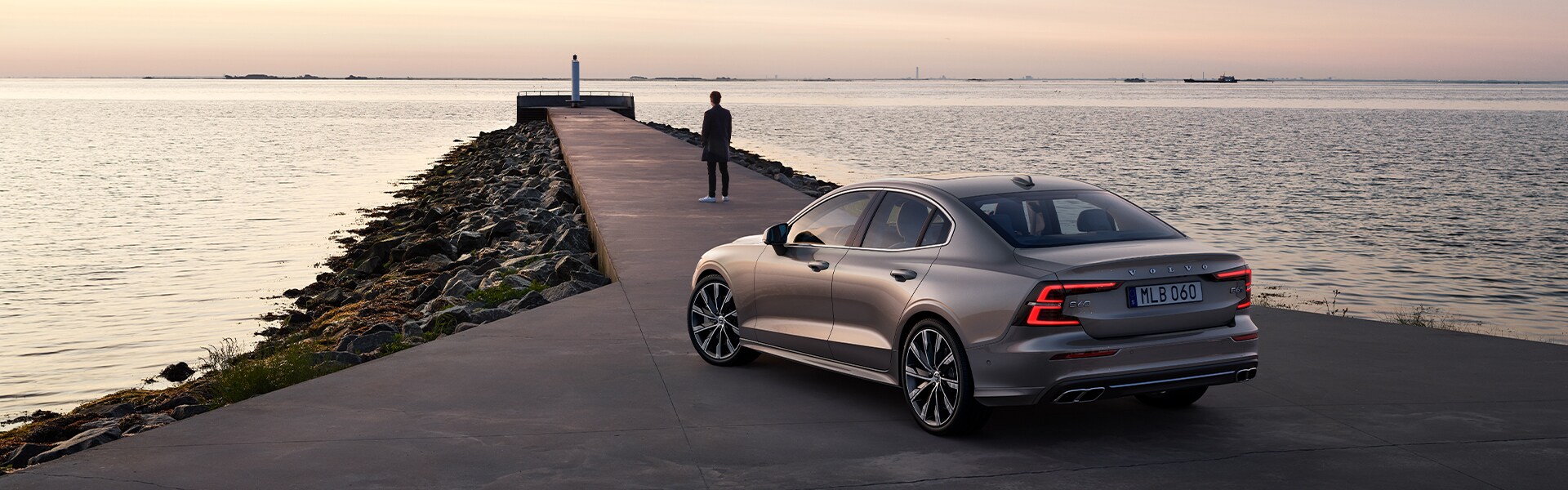 Volvo Cars New Bern is a Car Dealership near Pollocksville, NC | Silver 2021 Volvo S60 Parked In Front of Cement Pier Leading Out to Ocean