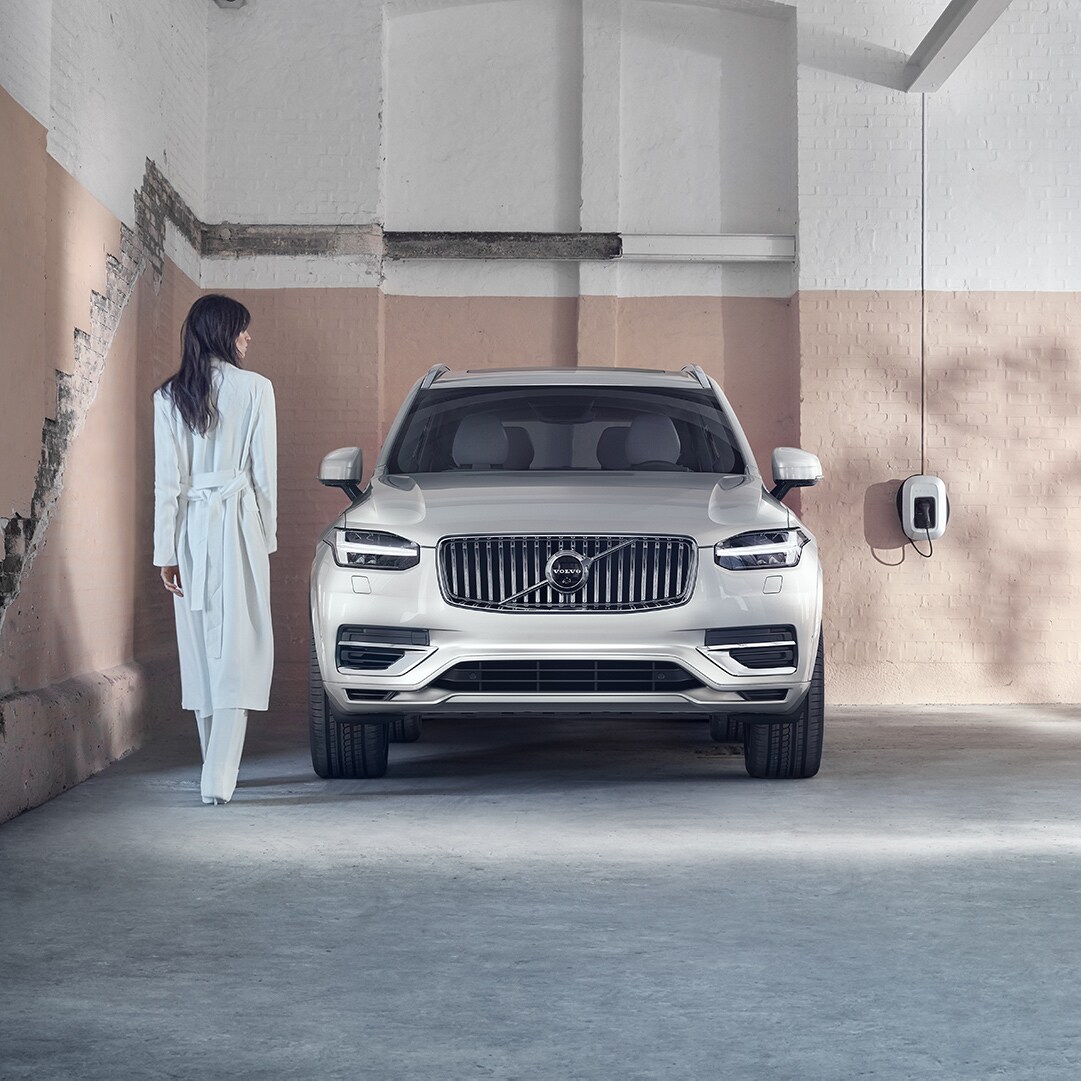 Volvo Cars New Bern is a Car Dealership near Oriental NC | Light Grey MY20 Volvo XC90 parked in garage with pink wall and woman dressed in all white near passenger door