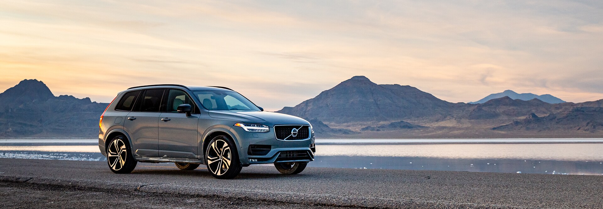 Volvo of New Bern is a Car Dealership in New Bern near Neuse Forest NC | Silver 2020 XC90 Driving by Lake and Mountains at Sunset Volvo of New Bern is a Car Dealership in New Bern near Neuse Forest NC | Silver 2020 XC90 Driving by Lake and Mountains at Sunset
