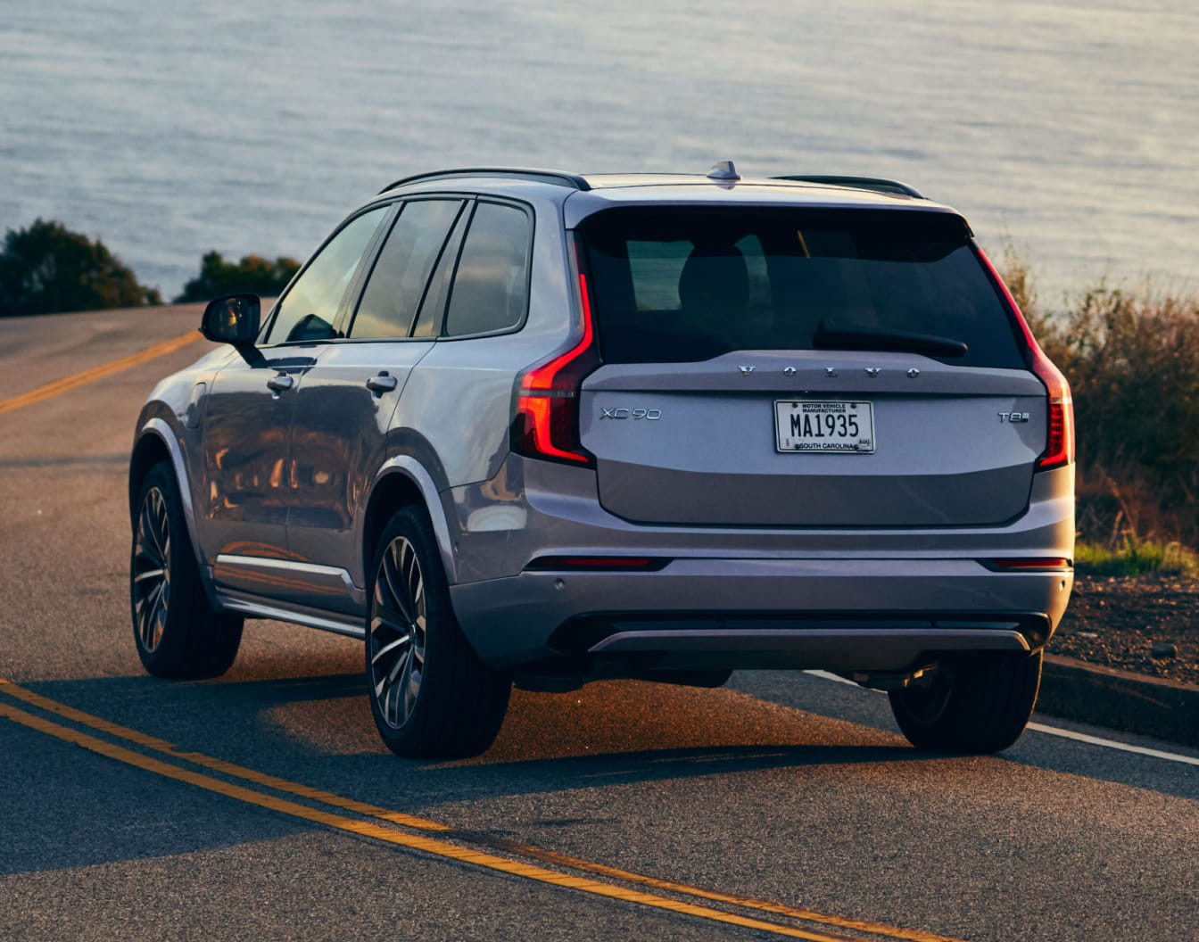 Volvo XC90 driving along a winding road beside a body of water, shown from a rear three-quarter angle.