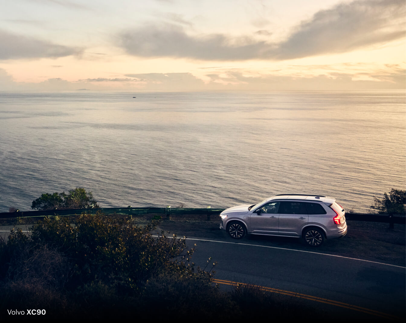 High-angle side view of silver Volvo XC90 driving on a coastal highway overlooking the ocean at sunset.