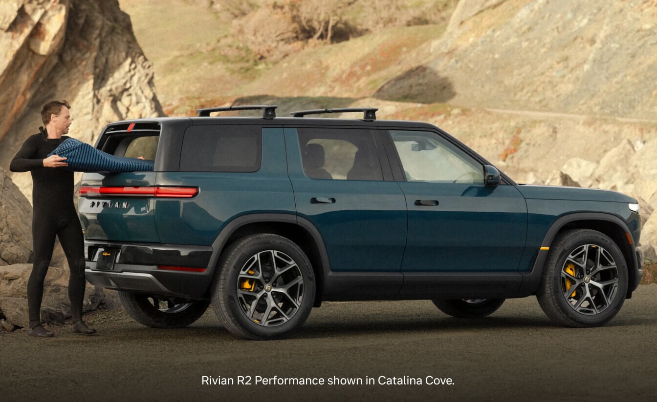 Rear three-quarter view of a 2026 Rivian R2 Performance in Catalina Cove parked along a rocky cliffside road.