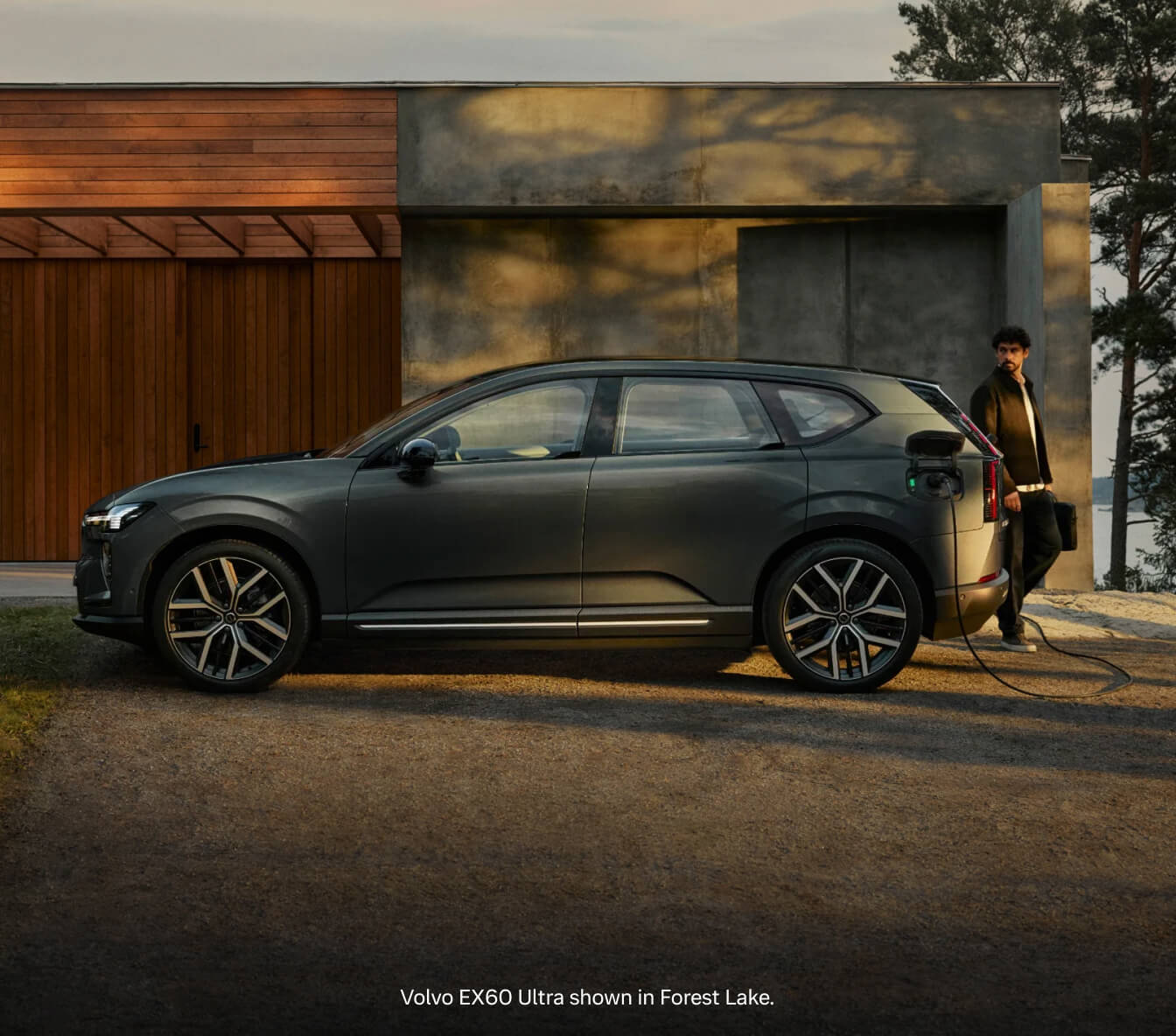 Side profile view of a 2026 Volvo EX60 Ultra in Forest Lake parked outside a contemporary home, charging at dusk.