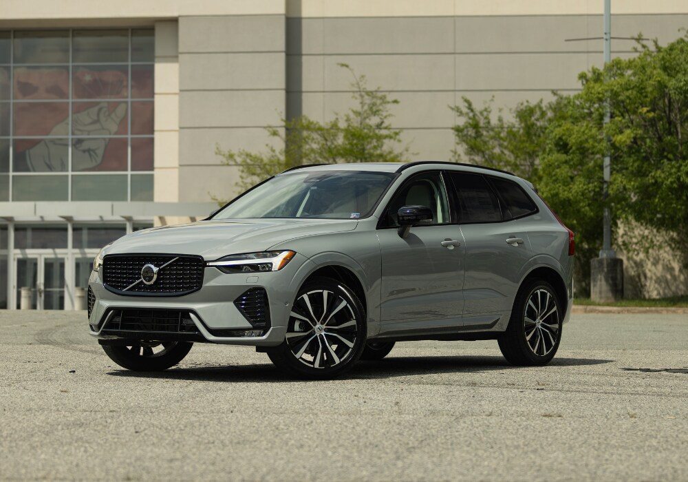 2025-volvo-xc60-for-sale.png Gray Volvo XC60 SUV parked in a lot with a large building in the background. The Volvo logo is visible on the front grille