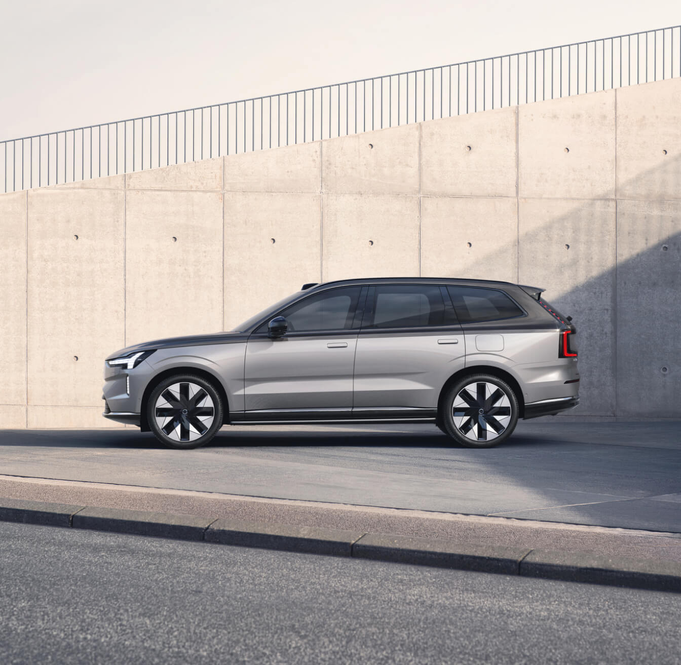 Side profile view of silver Volvo EX90 against a concrete wall background.