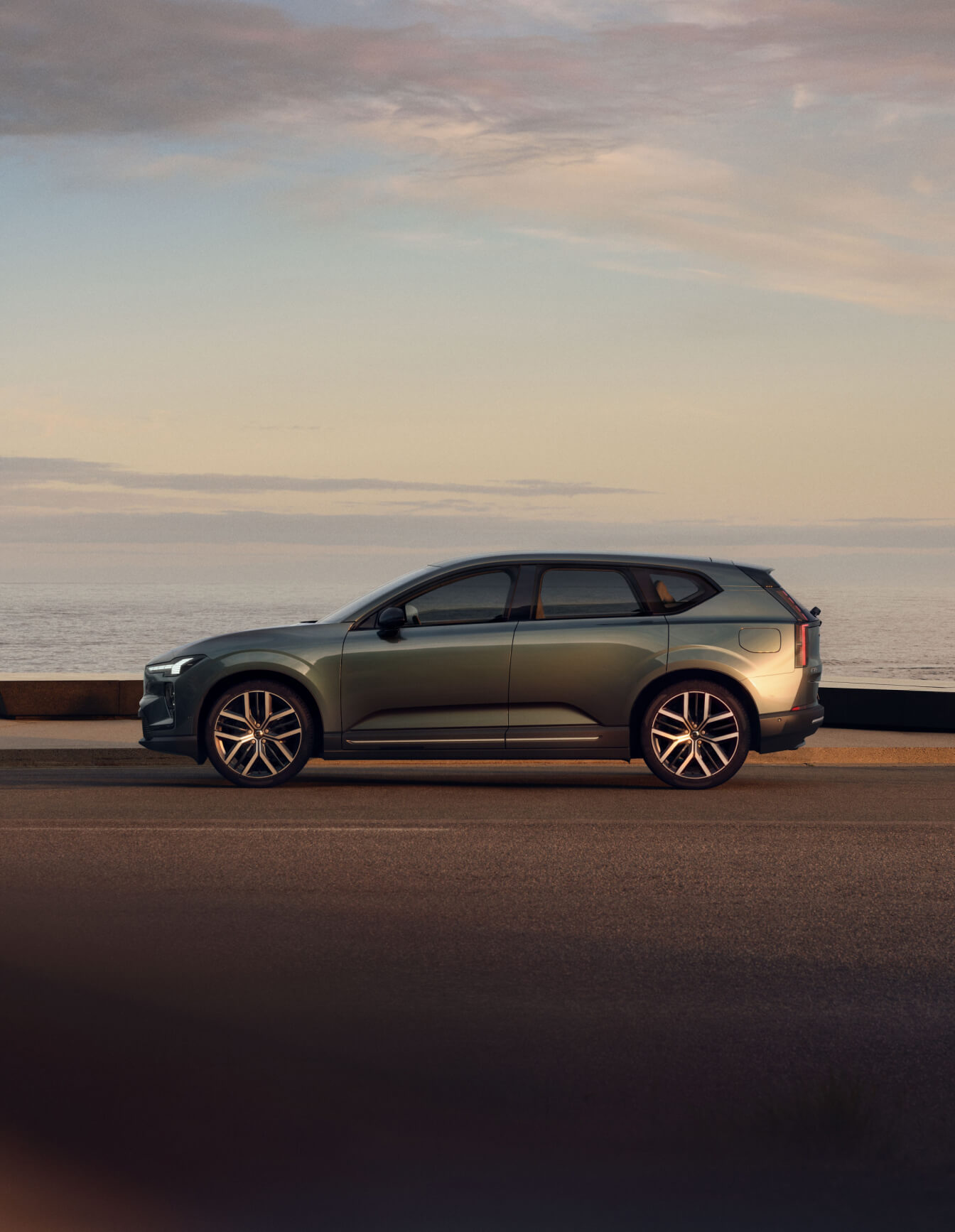 Side profile view of green Volvo EX60 parked on a coastal road overlooking the ocean at sunset.
