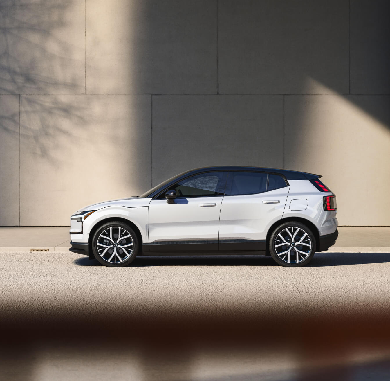 Side profile view of white Volvo EX30 with black roof against a concrete wall background.