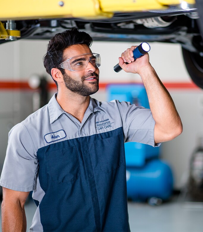 Best MPG At West Broad Hyundai in Henrico, VA | Mechanic in blue and grey uniform is inspecting the bottom of the car with a flash light and safety glasses
