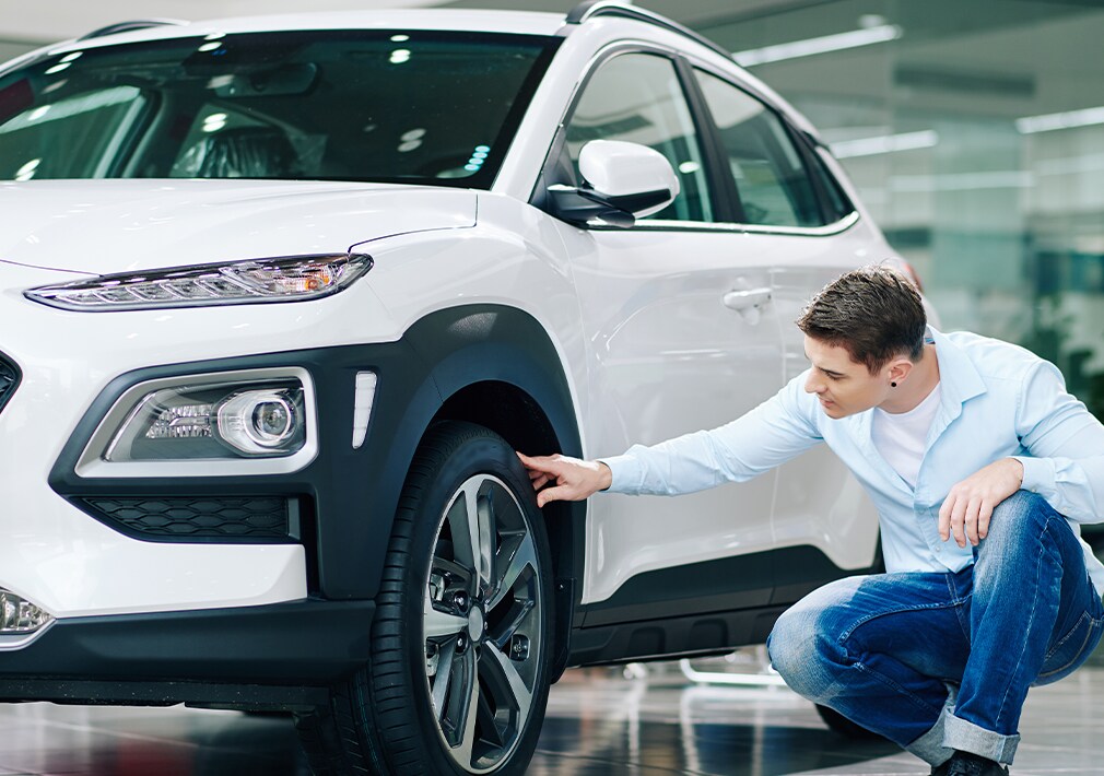 Learn Maintenance Warning Signs at West Broad Hyundai in Henrico, VA | Man in light blue button up shirt is checking the condition of his tires for his white Hyundai Kona