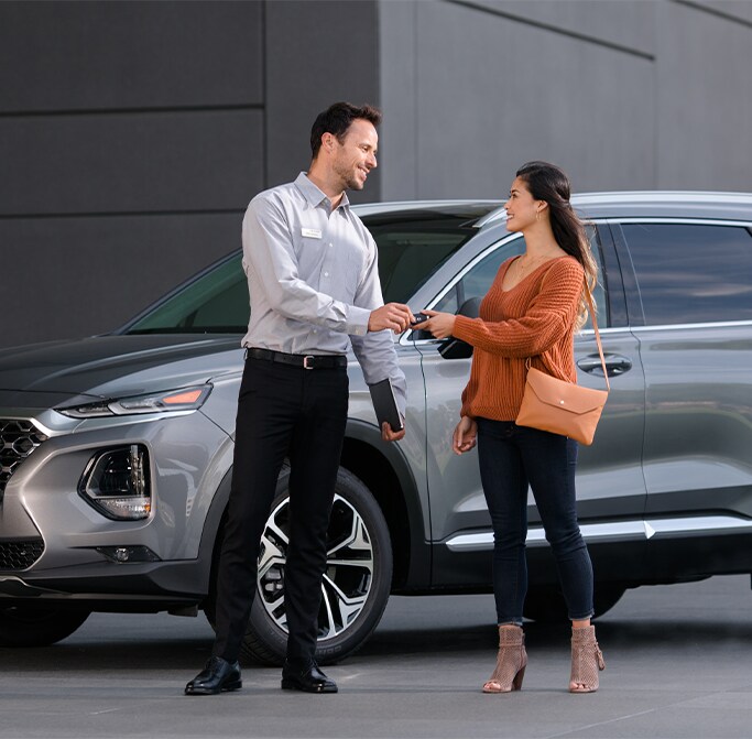Best MPG At West Broad Hyundai in Henrico, VA | Service adviser is handing over keys to a woman in orange sweater while a grey 2022 Hyundai Santa Fe is right behind them