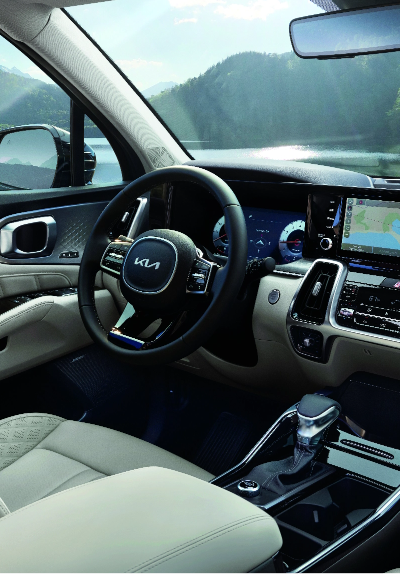 Luxury Kia SUV interior with cream leather seats, digital dashboard, and navigation display overlooking a scenic lake and forested mountains through the windshield