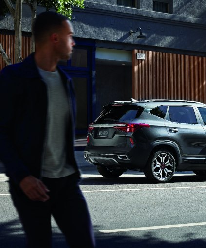Blurred pedestrian walking past a parked dark gray compact SUV on a city street in front of a modern building.