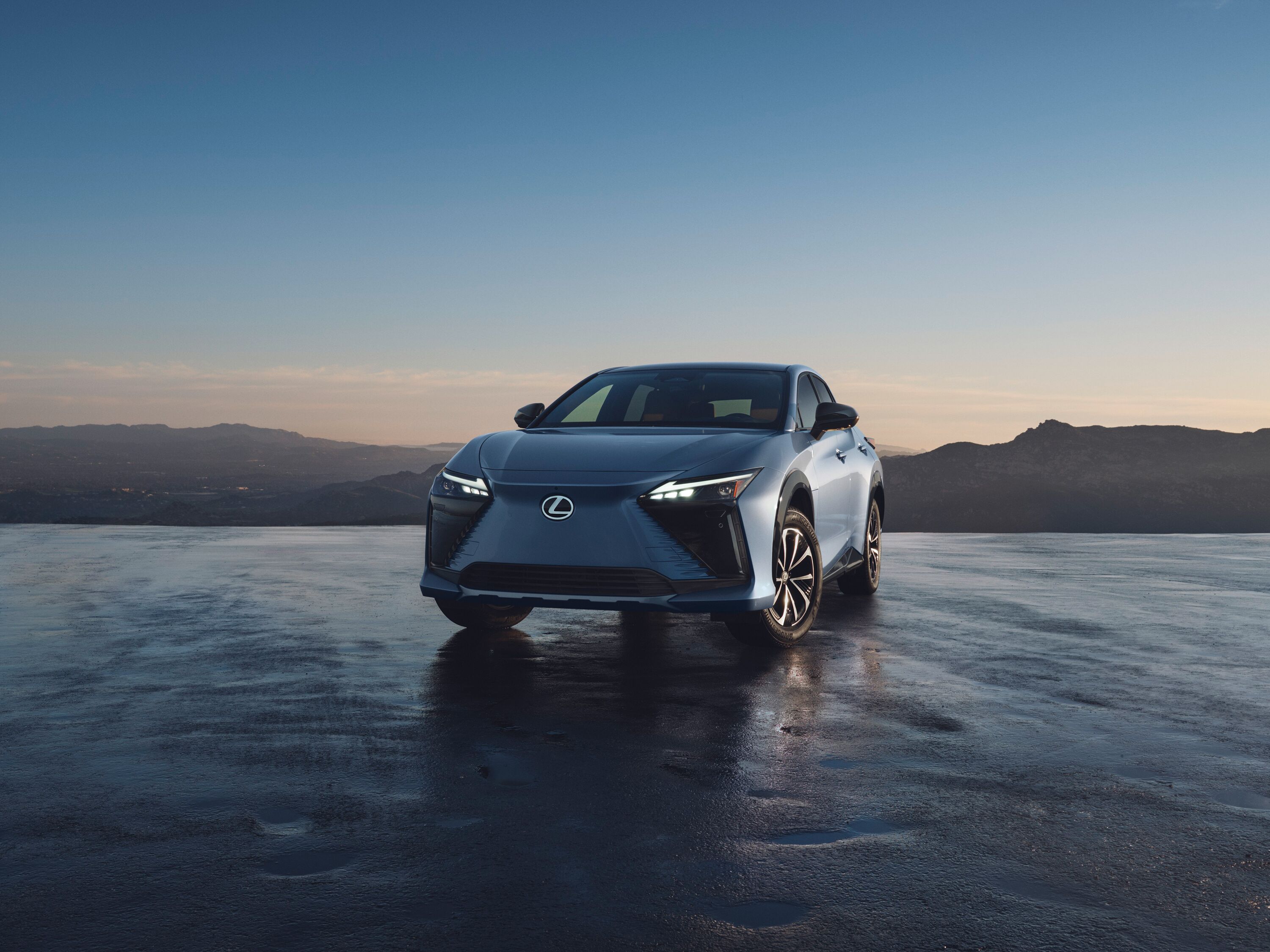 Which Lexus is right for me? Front view of an all-electric light blue Lexus RZ SUV parked on wet pavement with scenic mountain sunrise view.