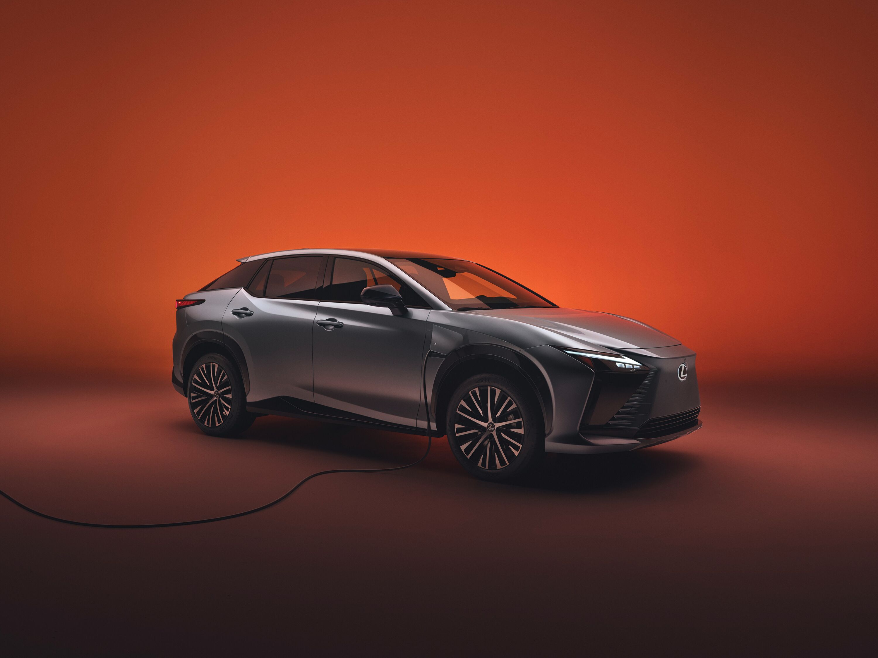 Which Lexus is right for me? Silver Lexus RZ all-electric luxury SUV connected to a charging cable against a warm orange studio background.