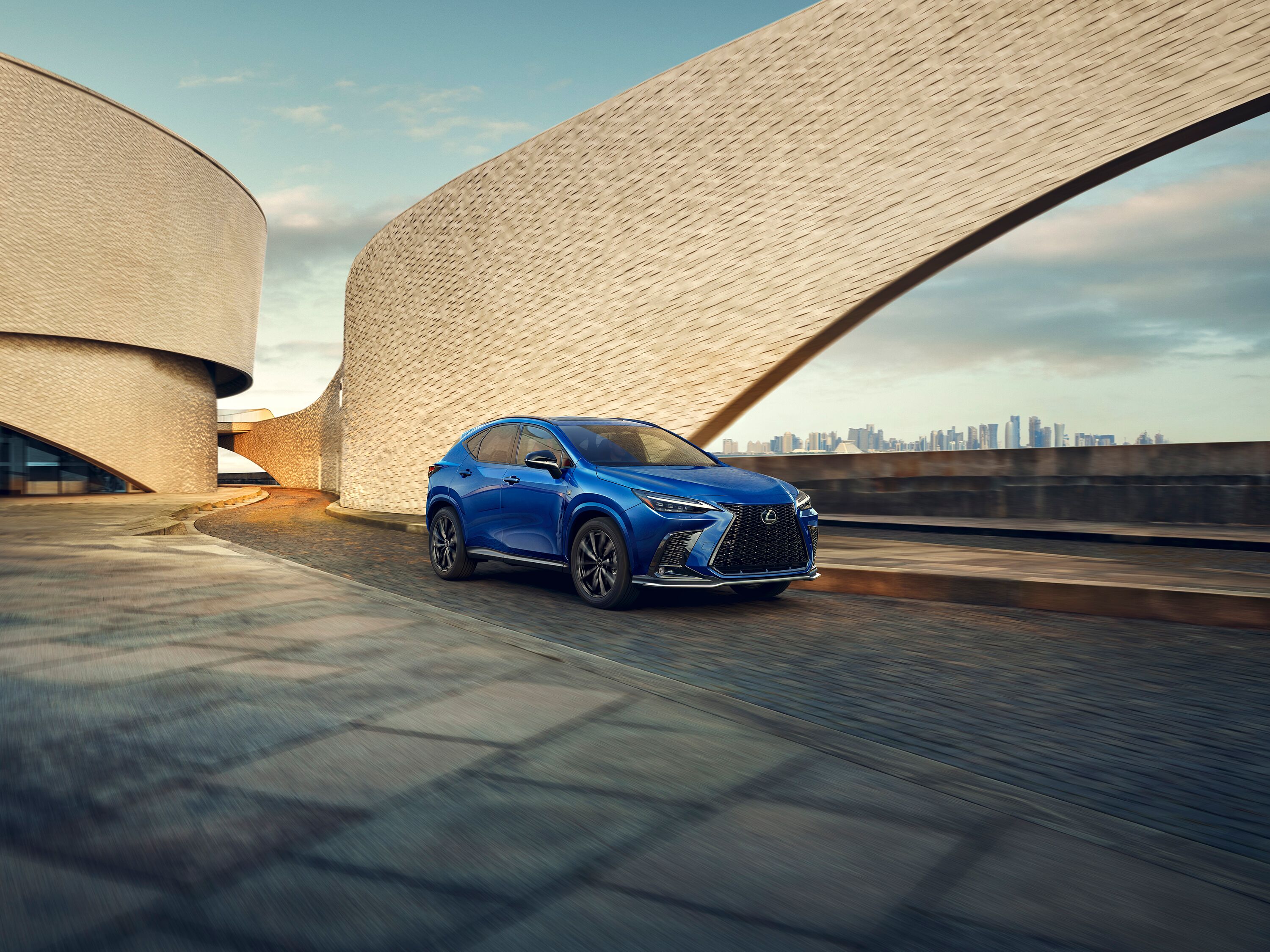 Which Lexus is right for me? Blue Lexus NX luxury compact SUV driving past modern architectural buildings at sunset.