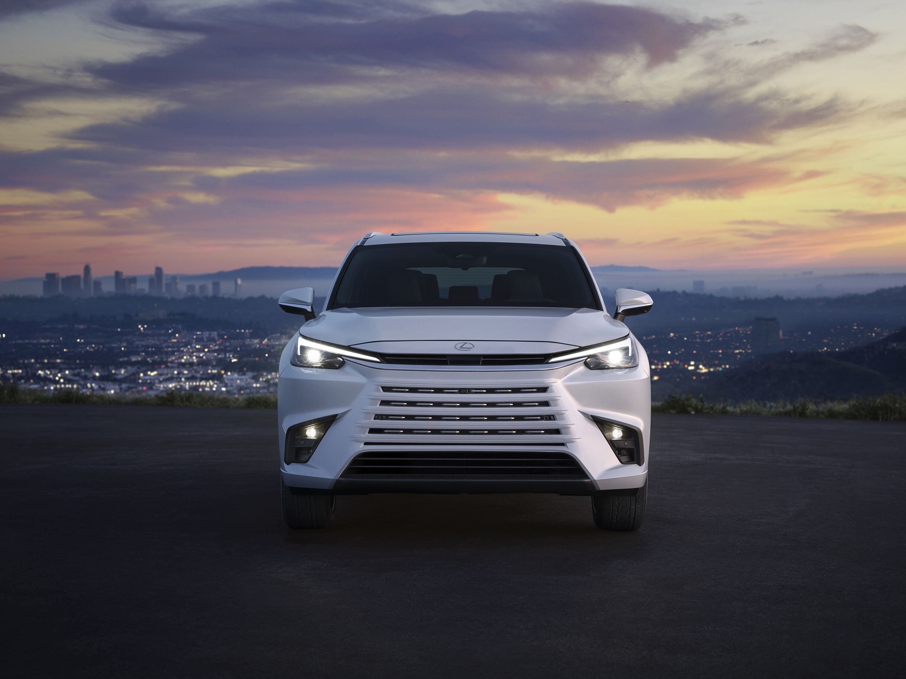 Which Lexus is right for me? Front view of a white Lexus TX three-row luxury SUV overlooking a city skyline at sunset.