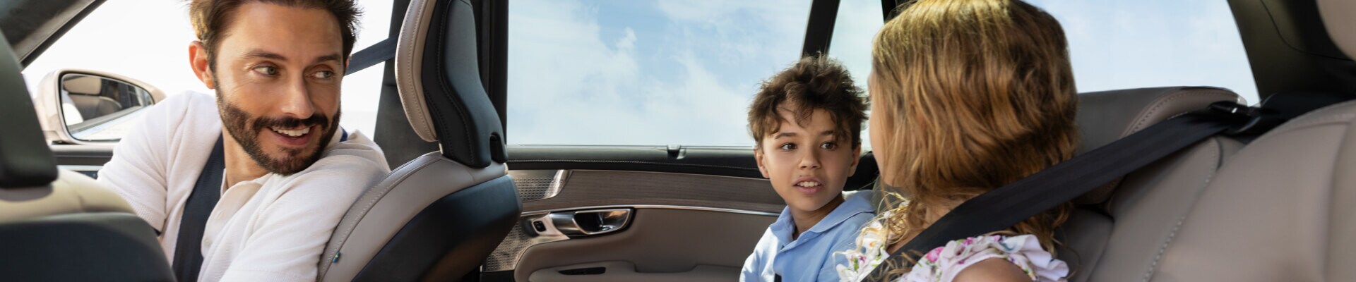 Interior view of a Volvo XC90. A man in the front passenger seat smiles while looking back at children in the rear seats.