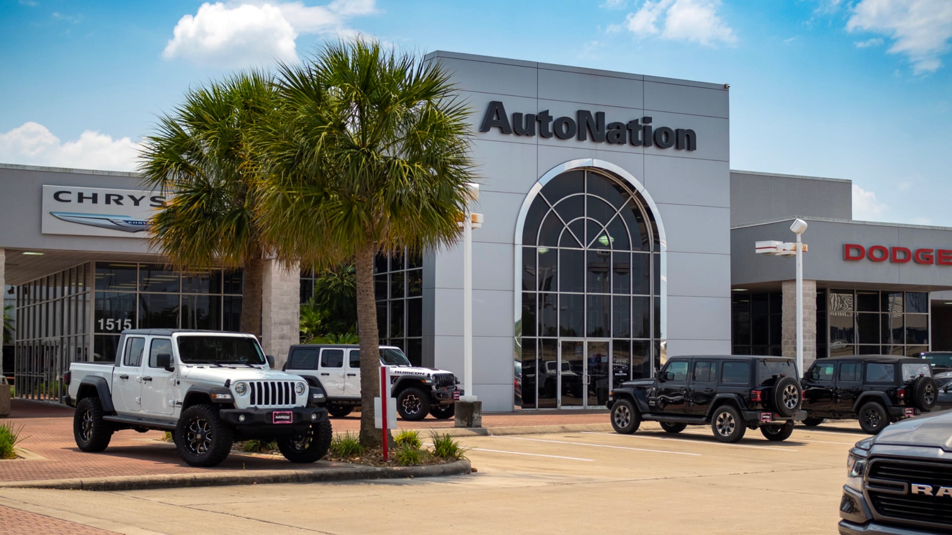 Exterior view of AutoNation Chrysler Dodge Jeep Ram Houston during the day. There is a clear blue sky and many vehicles parked near the building. Two palm trees are visible in the parking lot.