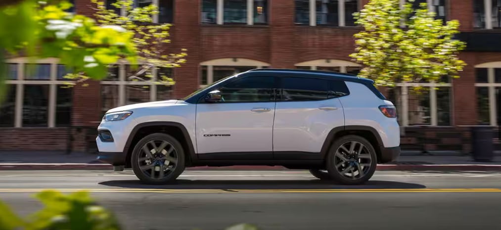 A white 2026 Jeep Compass driving through a city street near A.M. Maus & Son.