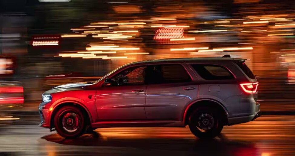 Side view of a silver 2026 Dodge Durango driving at night with blurred city lights, showcasing performance and style, available at A.M. Maus & Son.