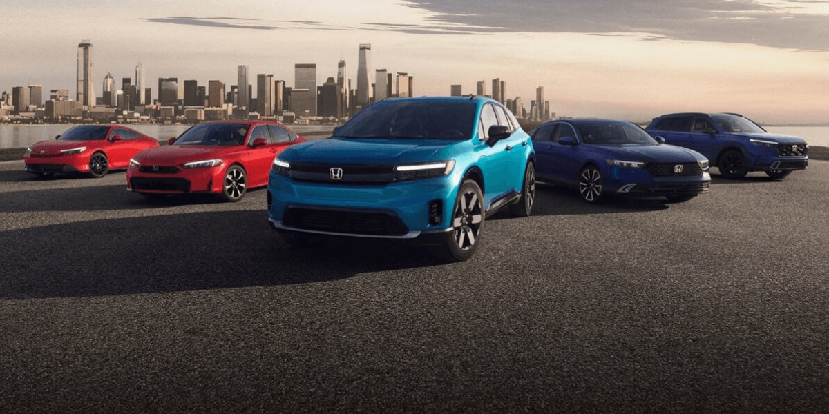 Five Honda vehicles in red and blue displayed together on a waterfront lot with a city skyline at dusk