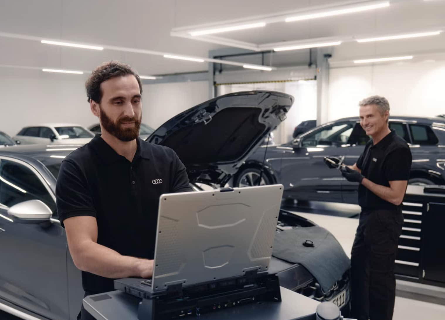 Audi service technician running computer diagnostics on a laptop while a second technician works on an Audi with the hood open in the Audi Annapolis service center