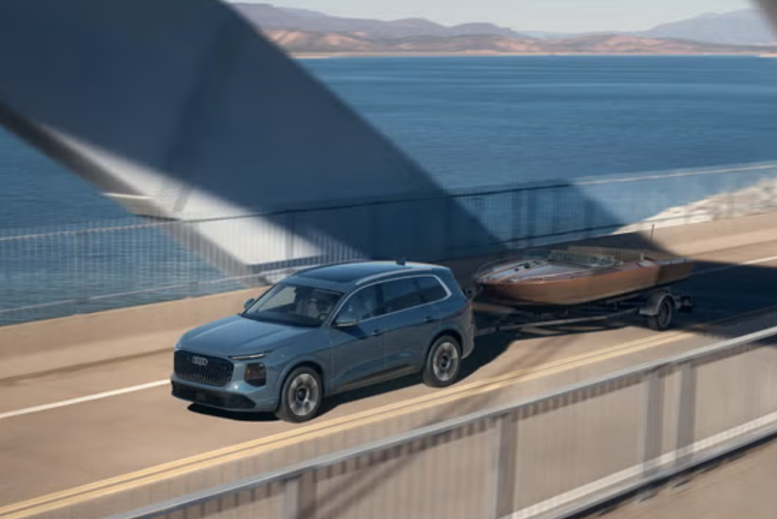 2027 Audi Q7 towing a wooden boat on a trailer across a bridge over the water