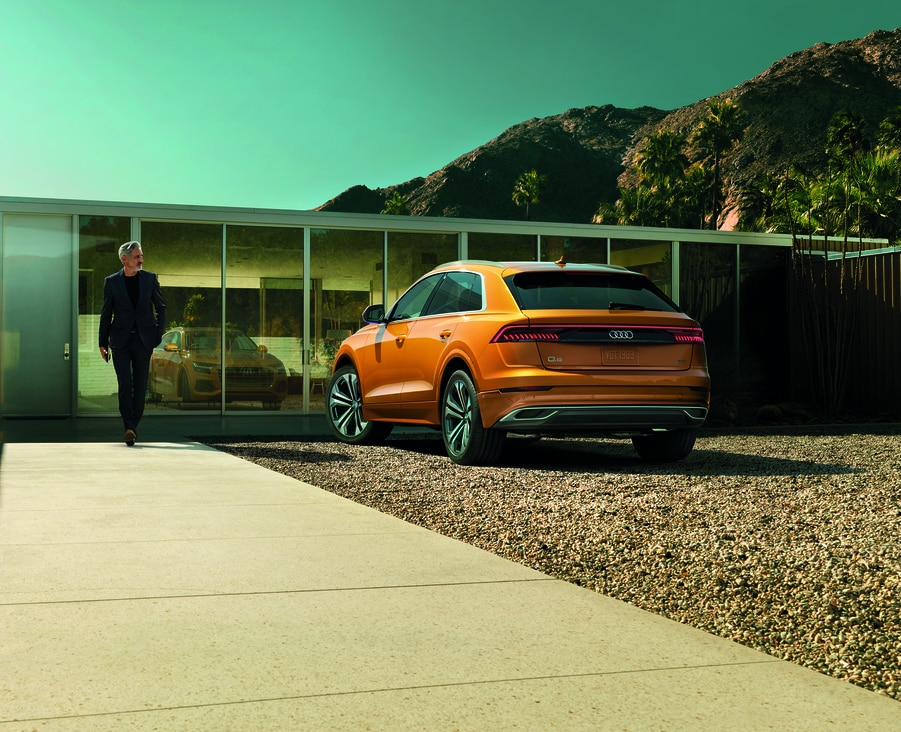orange Audi Q8 parked on the gravel next to a glass-covered modern home, with the driver walking towards it