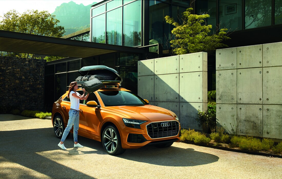 orange Audi Q8 SUV parked in the driveway of a modern home with the driver putting bags into a luggage storage rack on the roof