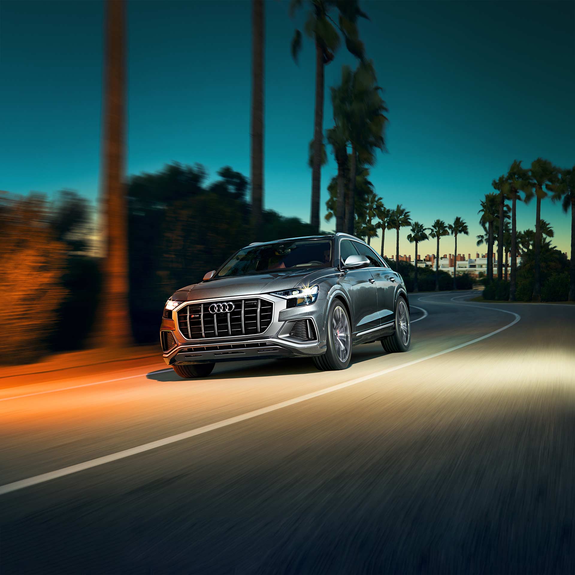 dark silver Audi Q8 SUV driving past a blurred palm tree lined street at sunset