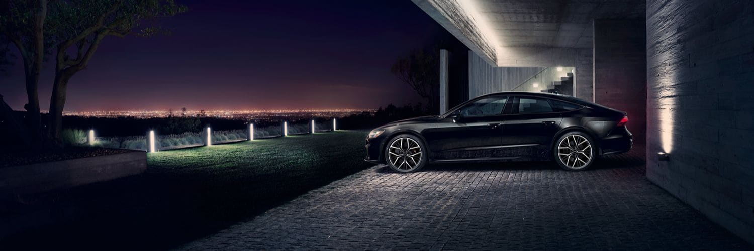 Black Audi S7 parked on stone driveway at night with city lights in the distance