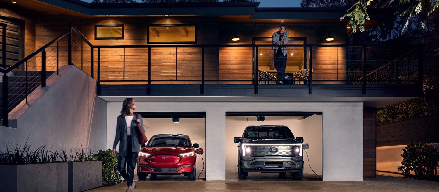 A 2022 Ford Mustang Mach-E and a Ford F-150 LightningTM are being 
charged in a garage at night while a man stands on a balcony looking at a
woman below, who is looking back at the man.